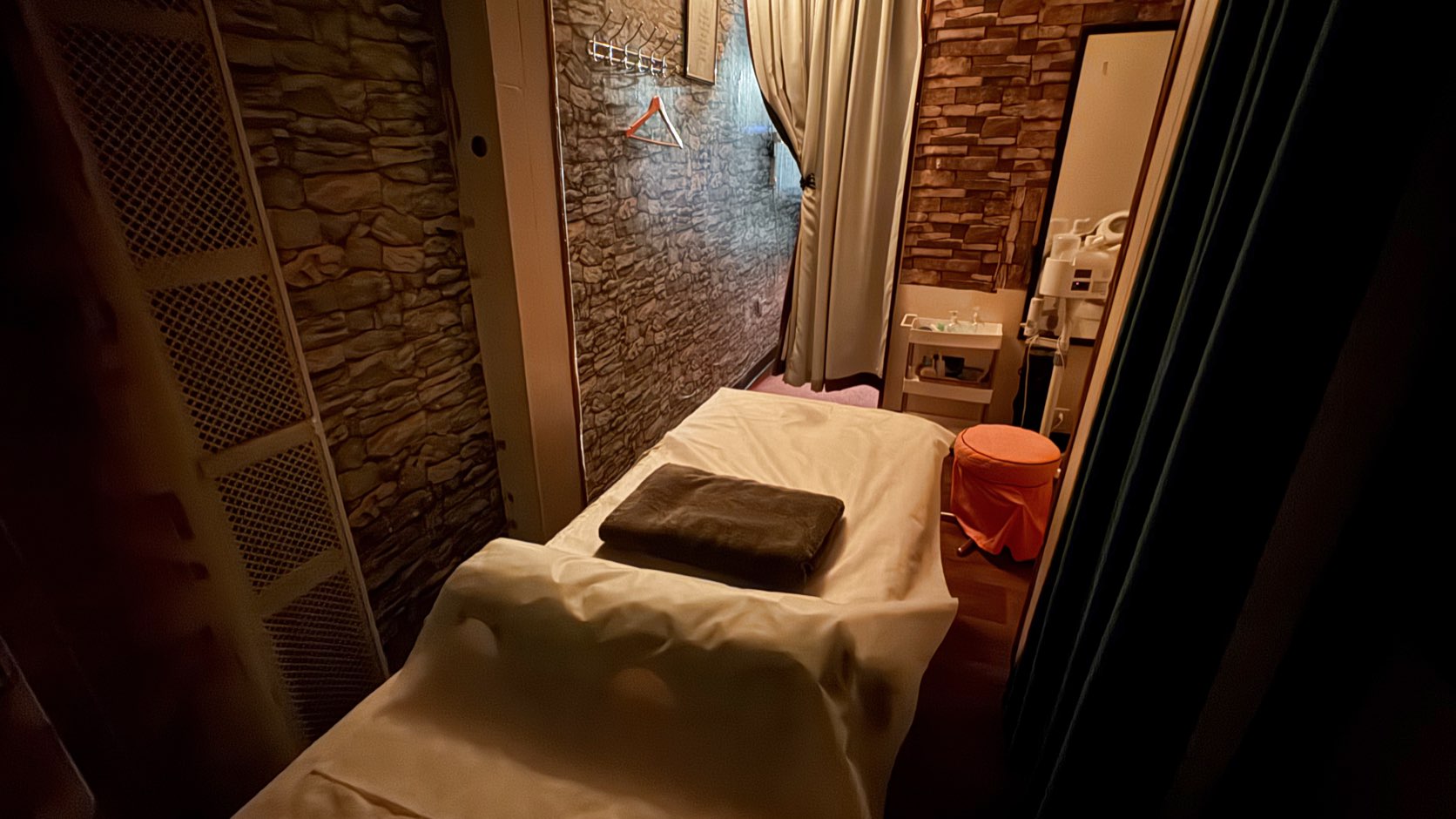 A treatment room at Elizabeth Spa: a massage table made up with fresh linen and a folded towel, faux-stone walls, a curtained doorway and a coat rail with one hanger.